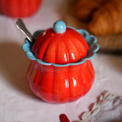 A glossy red ceramic pumpkin-shaped container with a matching lid, a scalloped light blue rim, and a small spoon resting inside, styled on a table with a blurred croissant in the background.