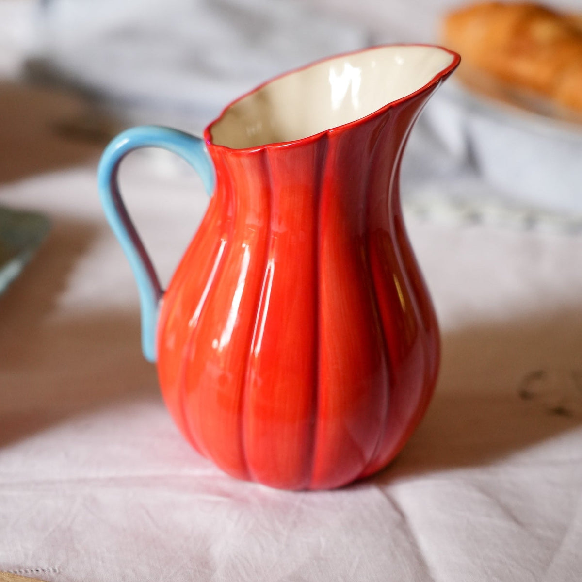 A glossy ceramic pitcher with a ribbed pumpkin-like body in vibrant red and orange tones, featuring a light blue handle and a cream-colored interior, set on a white tablecloth with a blurred kitchen background.