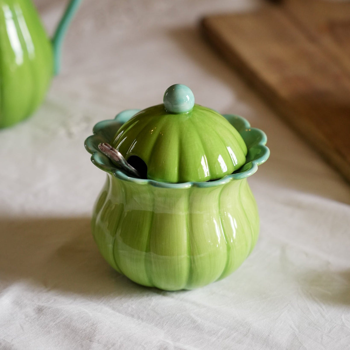 A green ceramic pumpkin-shaped container with a matching lid and small spoon, set on a white tablecloth with a wooden cutting board in the background.