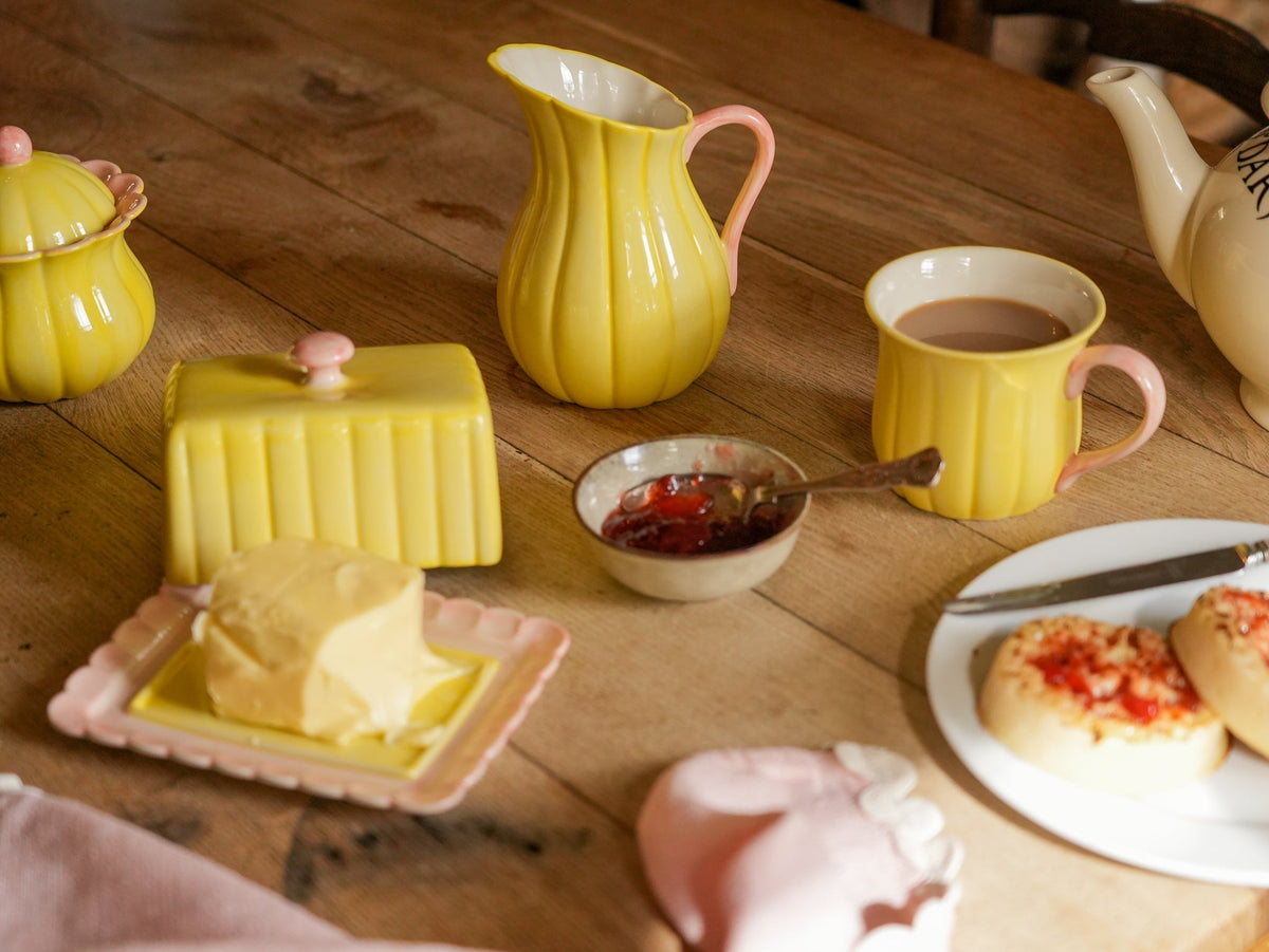 A cozy breakfast table scene featuring a coordinated yellow and pink ceramic tea and butter set, including a pitcher, mug, butter dish, and jam bowl, alongside scones and butter on a rustic wooden table.