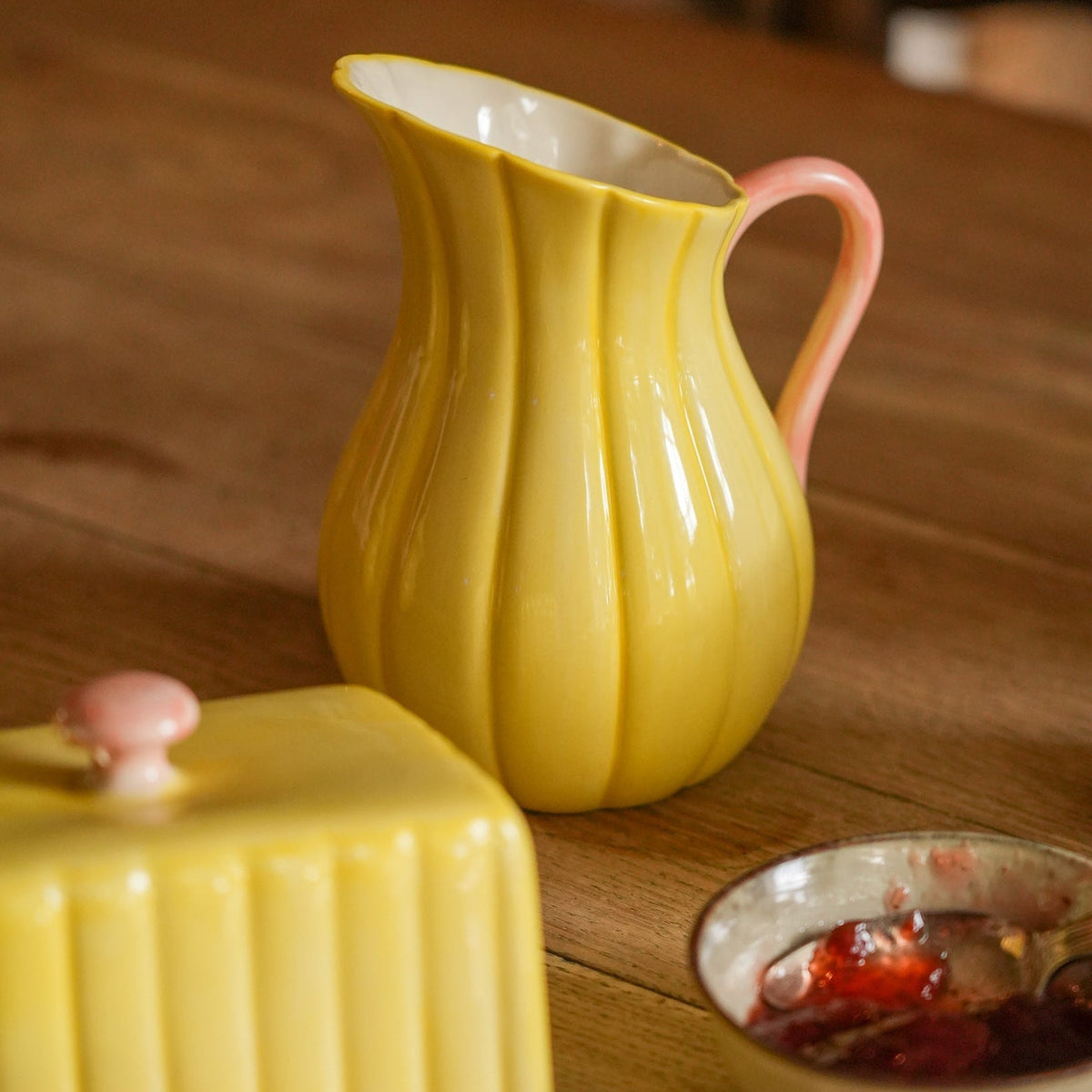 A yellow ribbed ceramic pitcher with a pink handle sits on a wooden table, alongside a matching yellow ribbed butter dish and a small bowl of jam.
