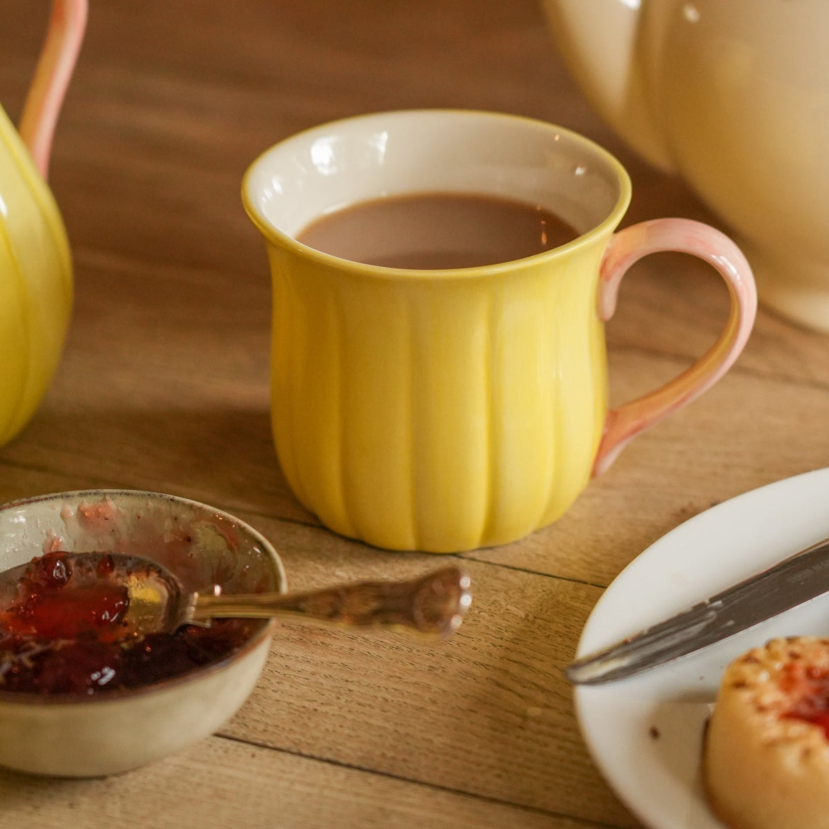 A yellow ribbed ceramic mug filled with tea sits on a wooden table, surrounded by a teapot, a small bowl of jam, and a plate with a pastry.