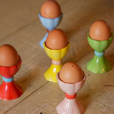 Five small ceramic egg cups in red, yellow, green, blue, and pink, each holding an brown egg, arranged on a rustic wooden tabletop.