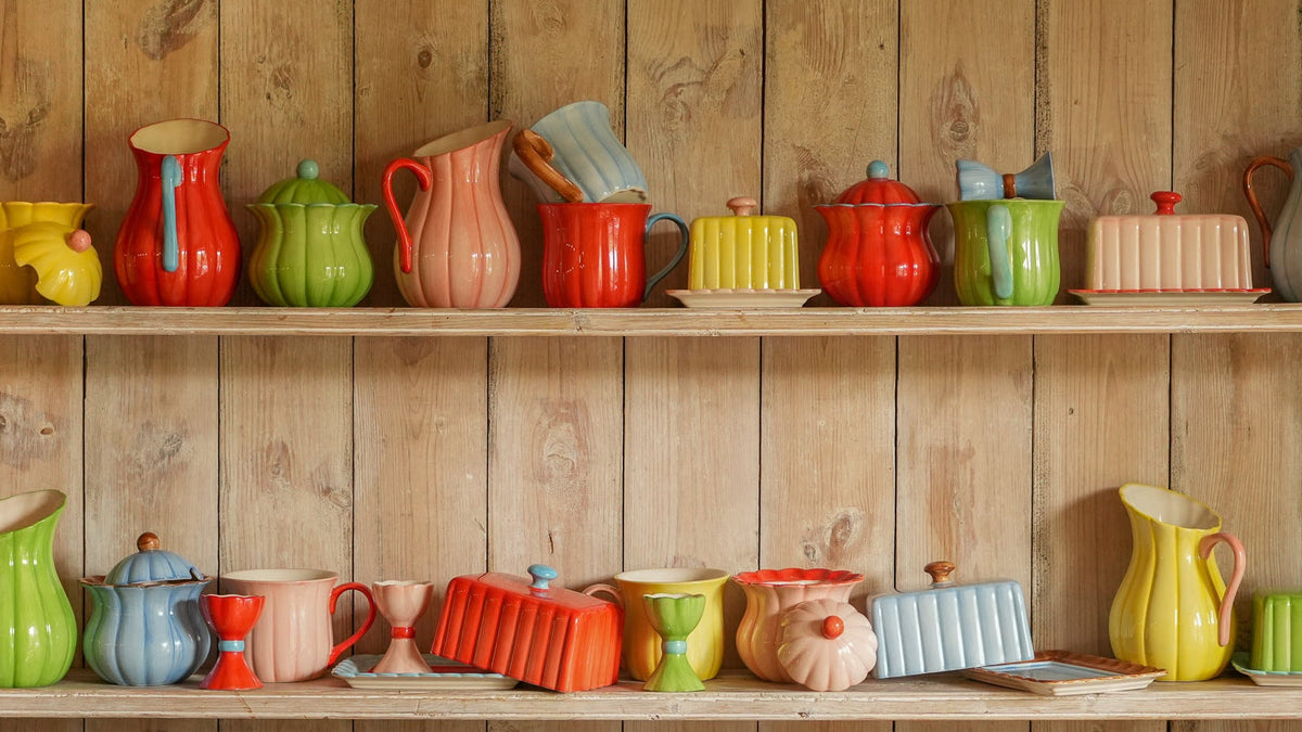 A collection of colorful ceramic kitchenware, including pitchers, mugs, and small pots, neatly arranged on two wooden shelves against a rustic wood-paneled wall.