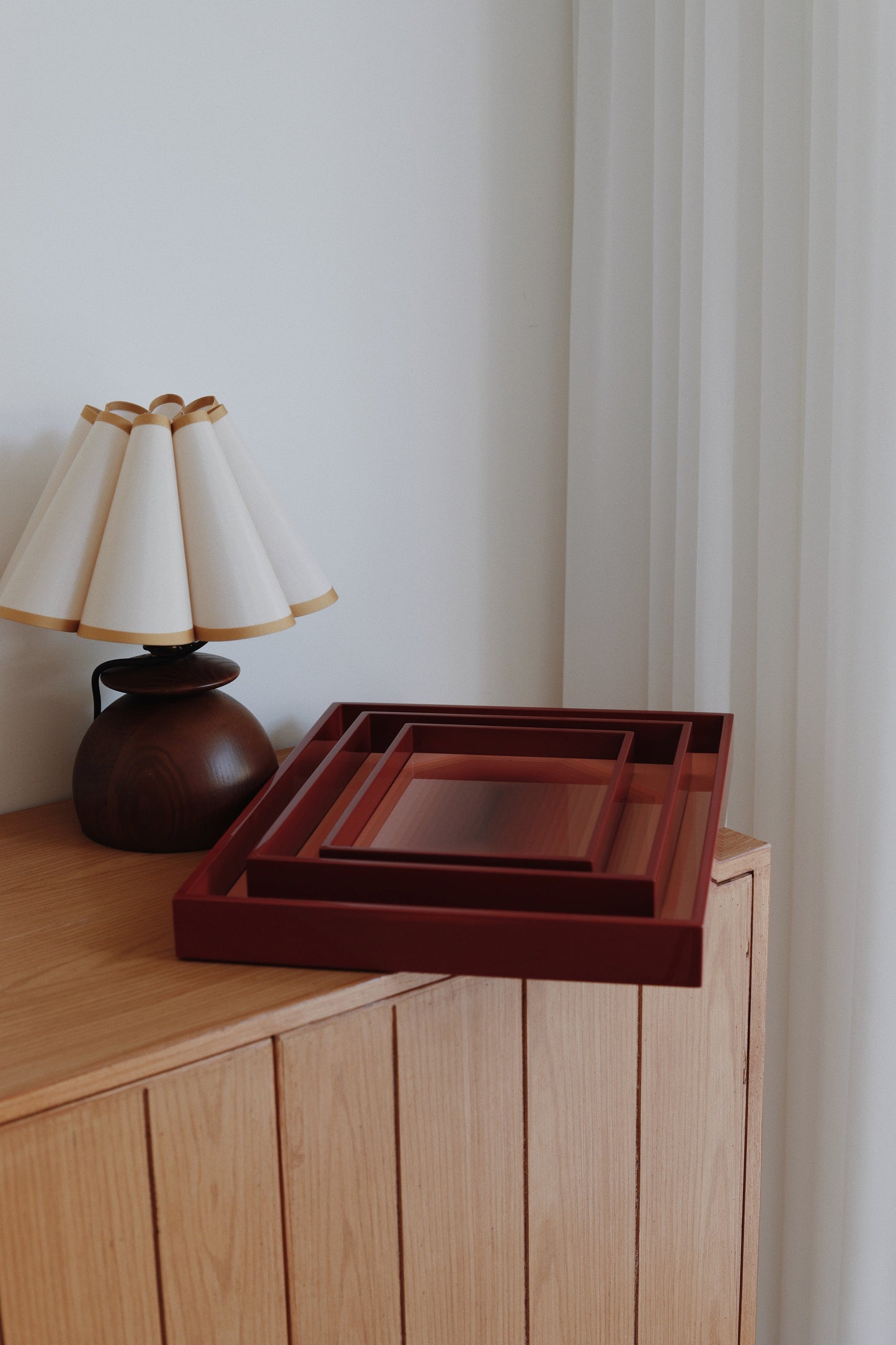 ested set of three deep red high-gloss lacquer trays resting on a natural oak sideboard next to a pleat-shade table lamp