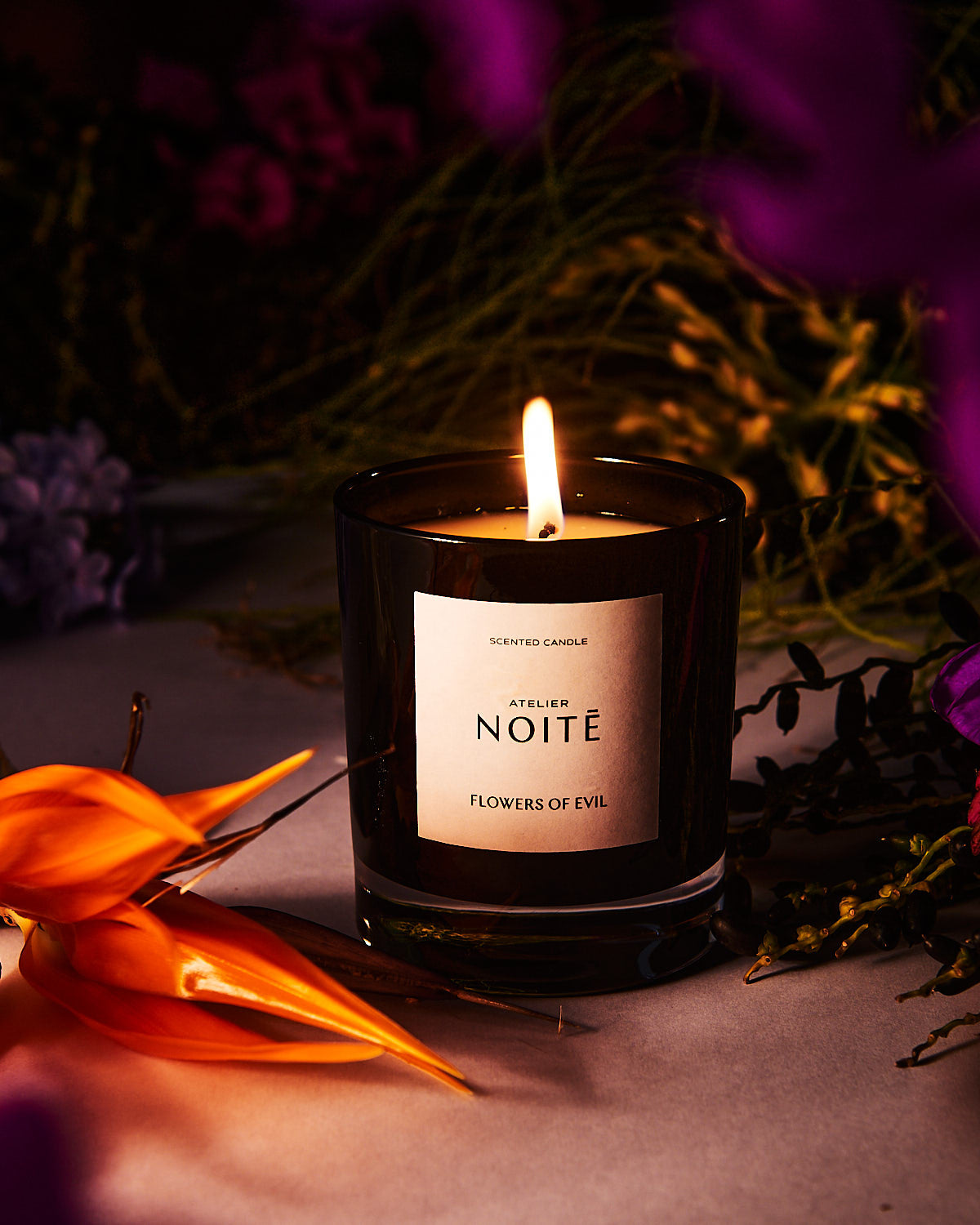 A candle in a black glass in dimly lit light with tropical flowers in the background
