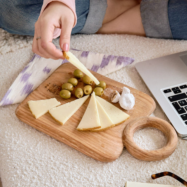 Person eating cheese and olives on a wooden board with a laptop and glasses in the background. - lifestyle