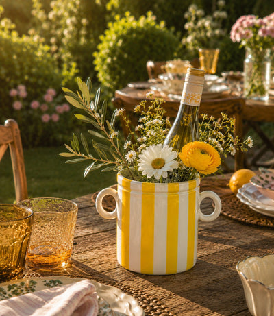 Decorative table setting with a yellow and white striped vase holding flowers and a bottle on a wooden table outdoors.