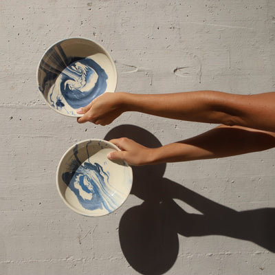 Two hands holding ceramic bowls with blue and white marbled design against a gray concrete wall. - lifestyle