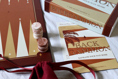 Backgammon board game set with a brown and gold board, two pink and white checkers, a branded box, play cards, and a red drawstring bag on a white fabric surface.
