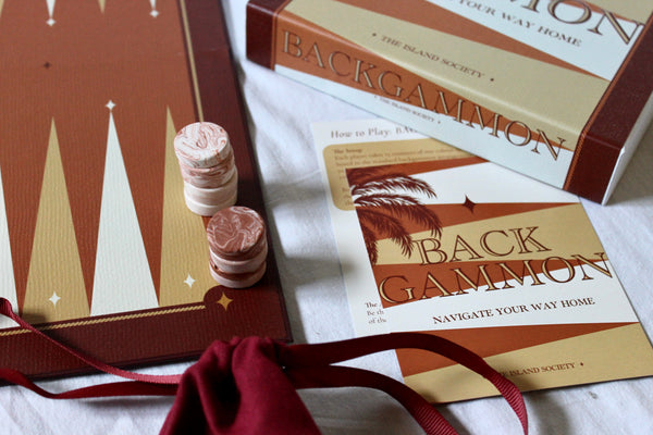 Backgammon board game set with a brown and gold board, two pink and white checkers, a branded box, play cards, and a red drawstring bag on a white fabric surface.