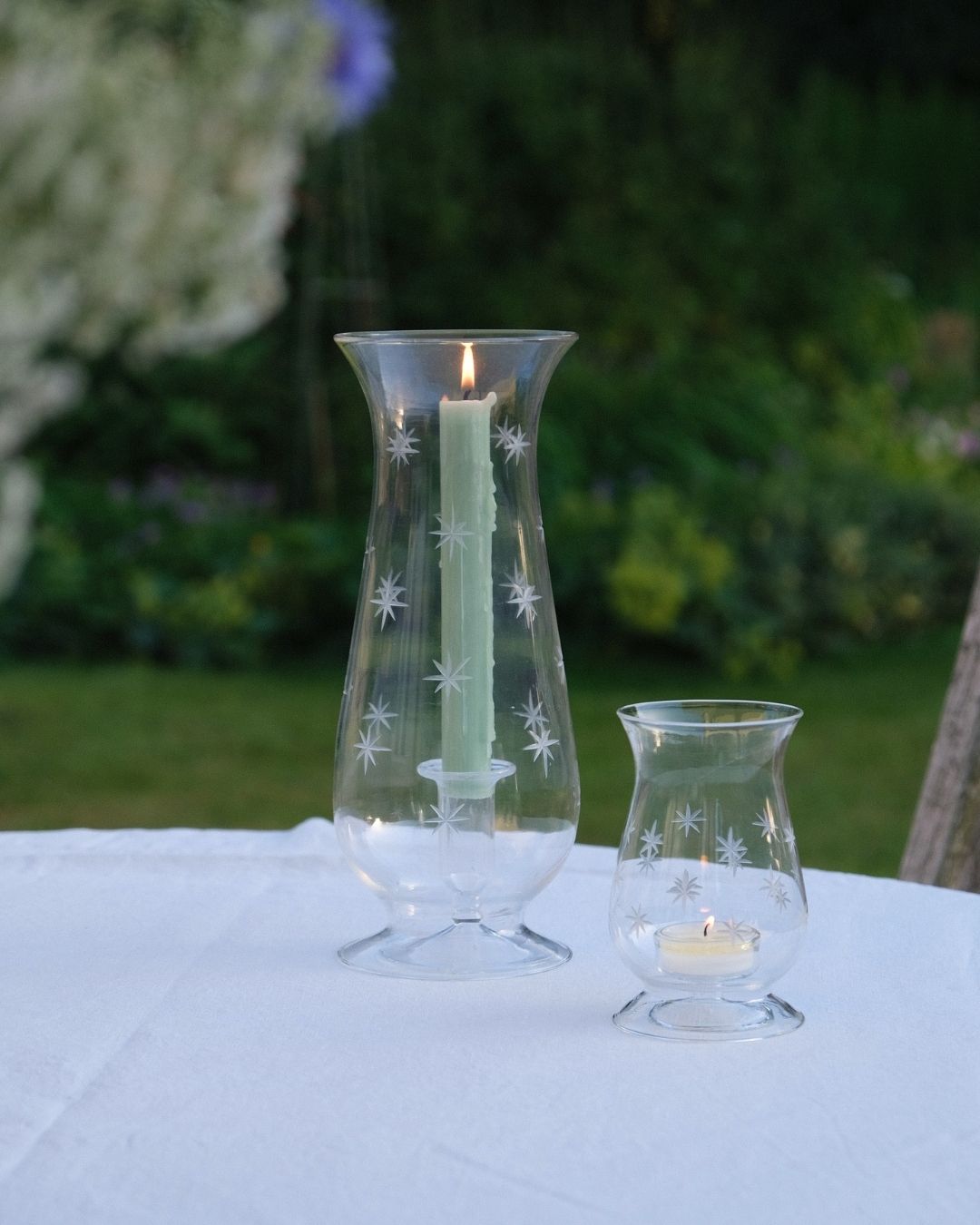 Two glass candle holders with lit candles on a white tablecloth outdoors. Issy Granger Hurricane Glass Lantern and Tealight holder
