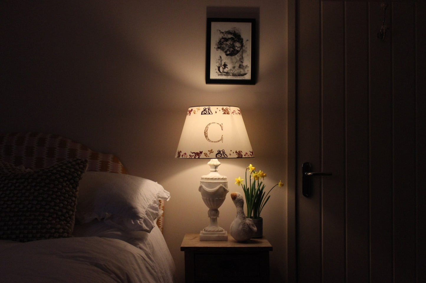 Personalised kids lampshade with woodland animals and letter C, glowing softly as part of cosy kids bedroom design, placed on a rustic wooden bedside table. The lamps is on giving the room a soft ,warm glow. A plush goose, daffodils, and white bedding add charm.