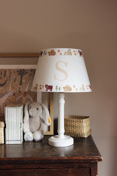 Personalised children’s table lamp with an initial “S” lampshade featuring woodland animal illustrations around the rim. The handmade nursery lampshade sits on a white lamp base on a wooden dresser beside books, a soft toy bunny and woven basket, creating warm, cosy children’s bedroom lighting.