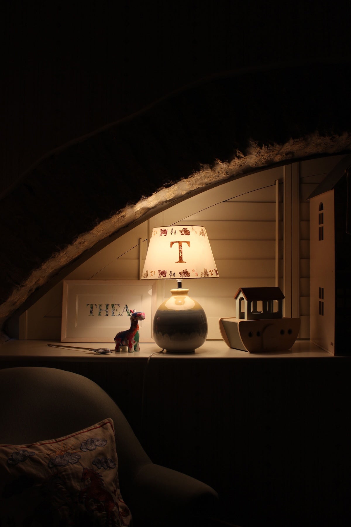 Children’s bedside table lamp with a personalised initial “T” lampshade, softly glowing in a cosy nursery setting. The handmade lampshade features small illustrated animal motifs around the rim, creating warm ambient lighting beside wooden toys and a dollhouse under a sloped ceiling.