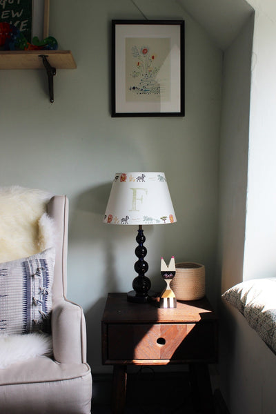 Safari lampshade with letter F, styled on side table in bright boys bedroom. Handmade safari lampshade for kids bedroom, personalised with letter F. Colourful empire light shade with safari animals, a green lampshade option for boys lampshade or kids bedroom design. The lamp is on a dark blue lamp base next to a cream arm chair with blue cushion and sheep skin. 