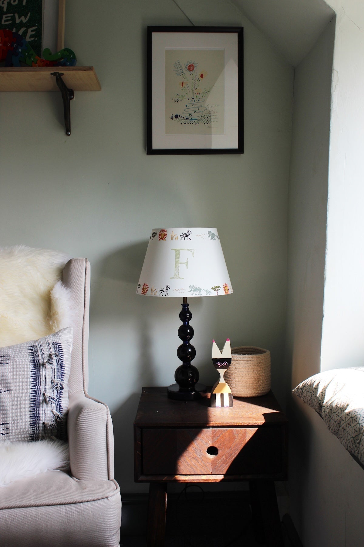 Safari lampshade with letter F, styled on side table in bright boys bedroom. Handmade safari lampshade for kids bedroom, personalised with letter F. Colourful empire light shade with safari animals, a green lampshade option for boys lampshade or kids bedroom design. The lamp is on a dark blue lamp base next to a cream arm chair with blue cushion and sheep skin. 