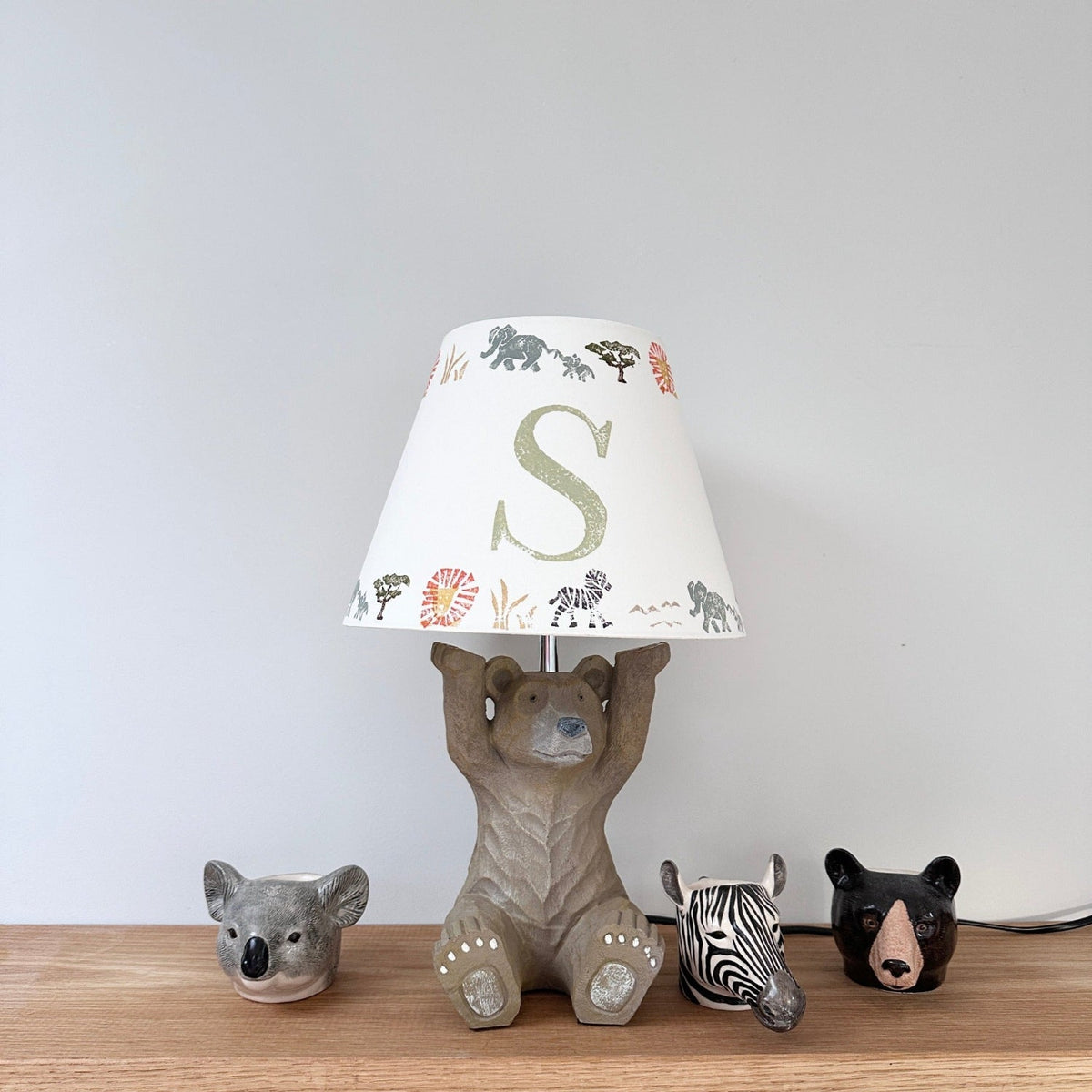 Personalised kids safari lampshade with letter S, colourful lampshade featuring elephants, zebras and lions. Empire lampshade design perfect for boys lampshade or kids lighting, ideal for kids bedroom decor, placed on a bear lamp base with animal egg cups.