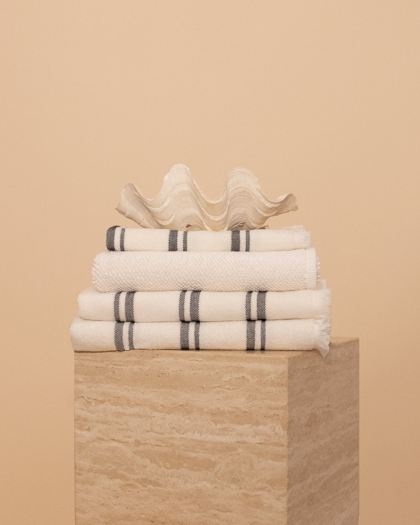 Kobn Crema Bathroom set includes towels, bathmat and handtowel, folded on a travertine plinth with a clam shell on top. 