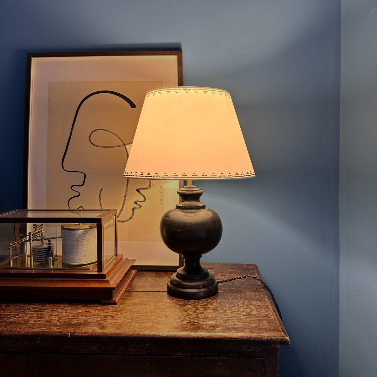 Romantic evening setting with a glowing cream empire lampshade featuring a lined triangle border in dark ink blue. The geometric lampshade casts a warm light, styled on a dark wooden lamp base against a deep blue wall.