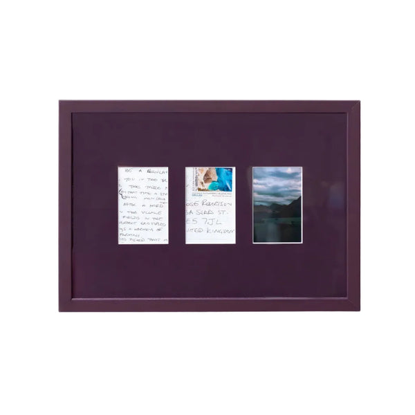 A square dark purple framed art piece mounted on a white wall, featuring three small vertical printed inserts with handwritten notes and a landscape photograph.
