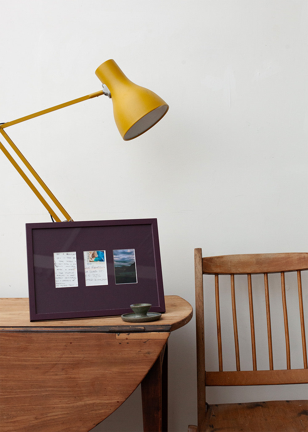 A dark purple picture frame with three small printed cards or photos inside, styled on a wooden table next to a yellow desk lamp and a wooden chair against a white wall.