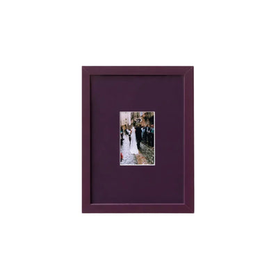 A square dark purple picture frame with a matching mat, featuring a small central photograph of a bride and groom, displayed on a clean white wall.
