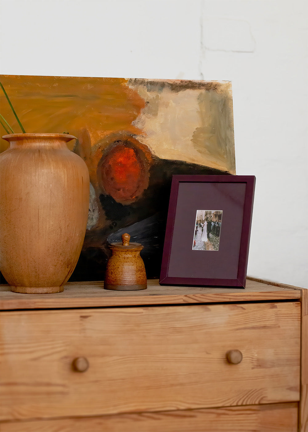 A styled interior scene showing a wooden dresser with a large terracotta vase, a small ceramic jar, a framed photo, and an abstract painting leaning against a white wall.