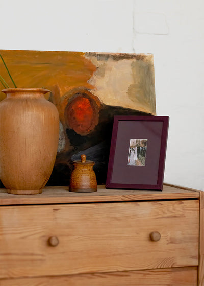 A styled interior scene showing a wooden dresser with a large terracotta vase, a small ceramic jar, a framed photo, and an abstract painting leaning against a white wall.