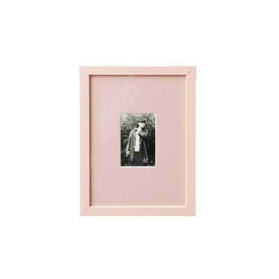 A vertical light pink picture frame with a white mat, showcasing a small black and white portrait photograph, mounted on a clean white wall.