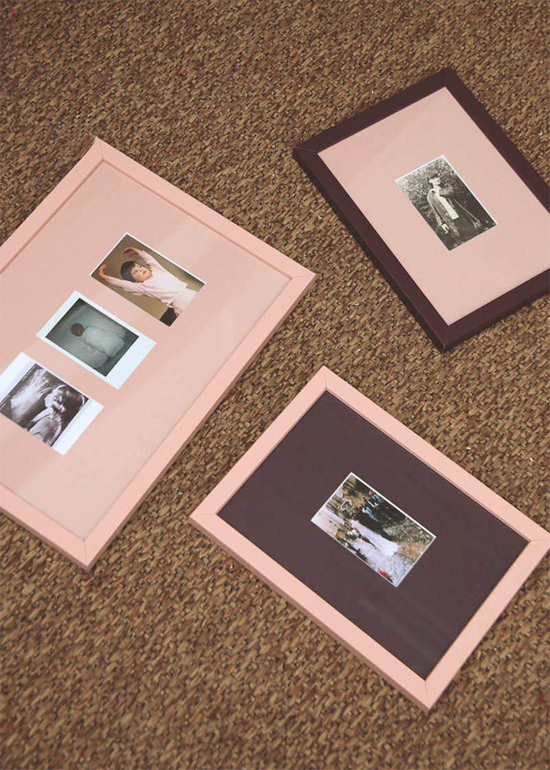 Three rectangular picture frames with pale pink borders and burgundy inner mats are arranged on a brown carpeted floor. 