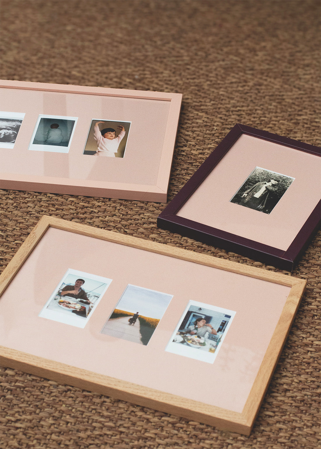 Three rectangular picture frames with pastel pink mats are arranged on a brown woven rug. One frame is light wood, one is dark wood with a deep purple border, and one is pale pink.