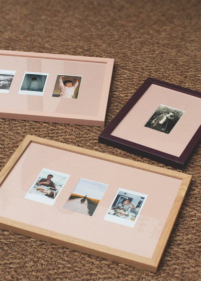 Three rectangular picture frames with pastel pink mats are arranged on a brown woven rug. One frame is light wood, one is dark wood with a deep purple border, and one is pale pink.