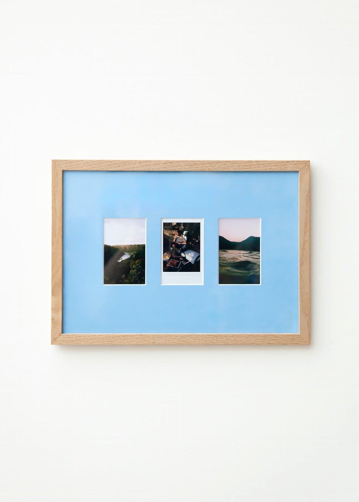A light wood framed wall art piece with a Sky blue mount and three small vertical landscape photographs arranged side by side, mounted on a plain white wall.