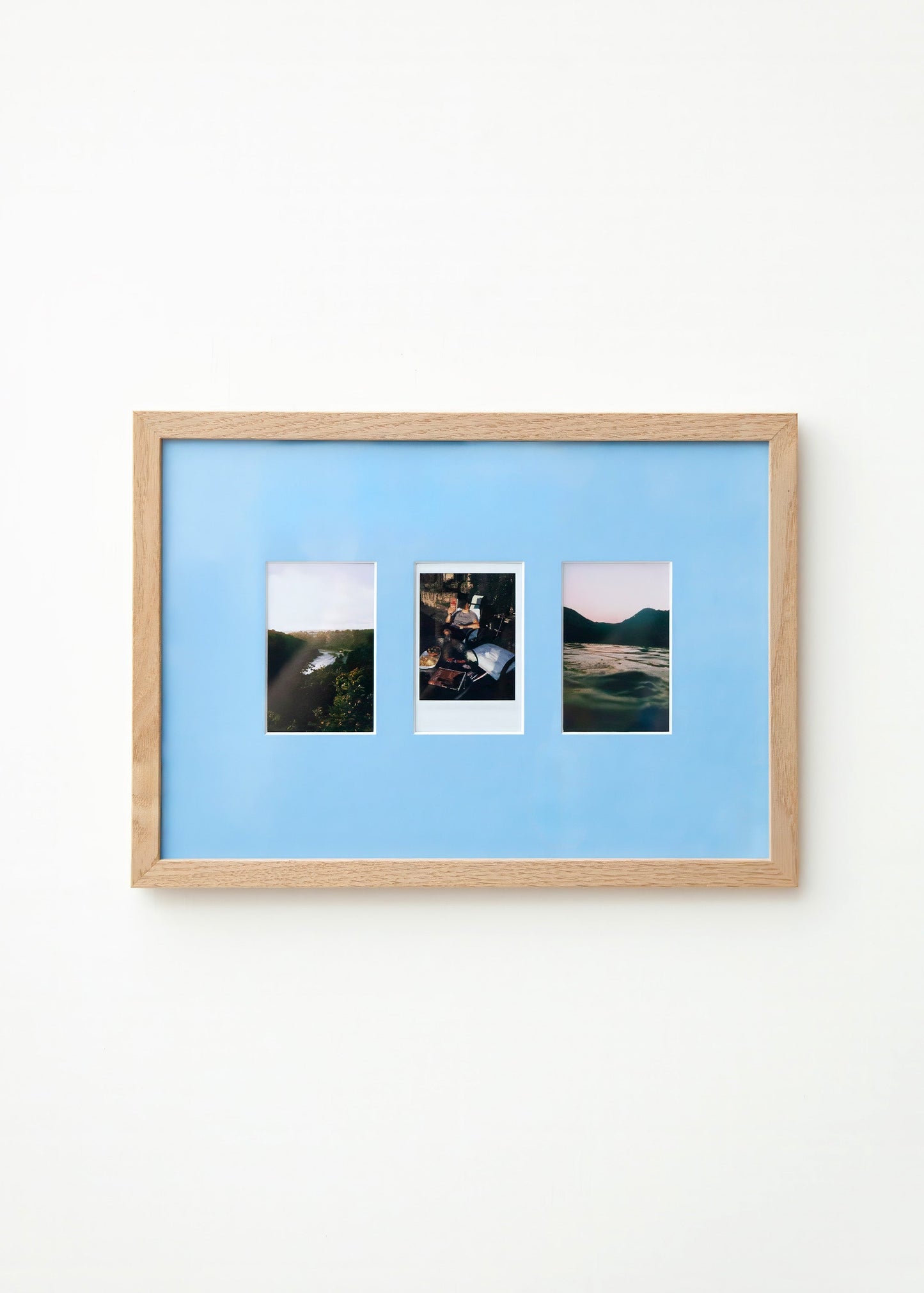 A light wood framed wall art piece with a Sky blue mount and three small vertical landscape photographs arranged side by side, mounted on a plain white wall.