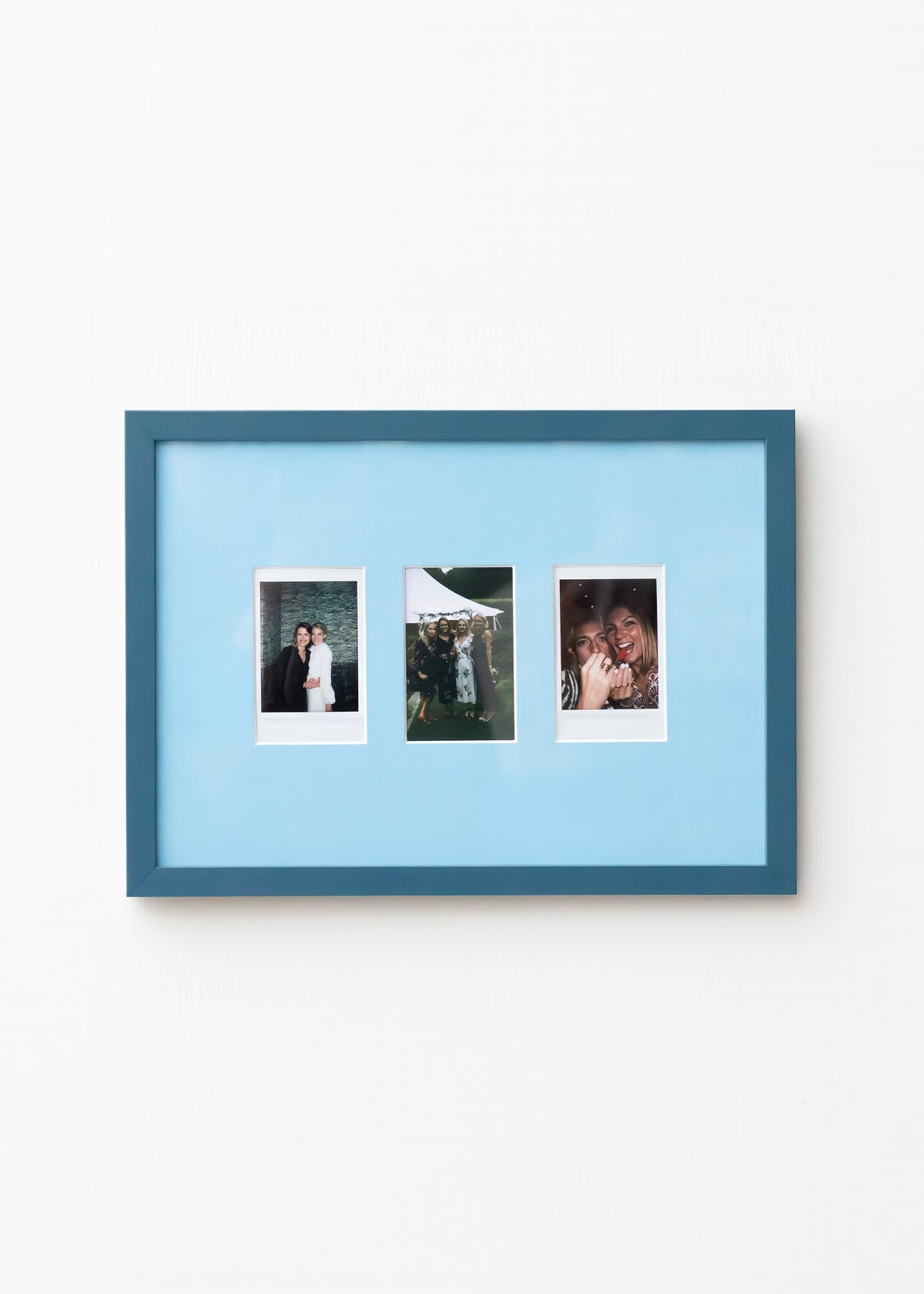 A blue picture frame with a light blue mat, holding three vertical instant-style photographs, shown hanging on a plain white wall.