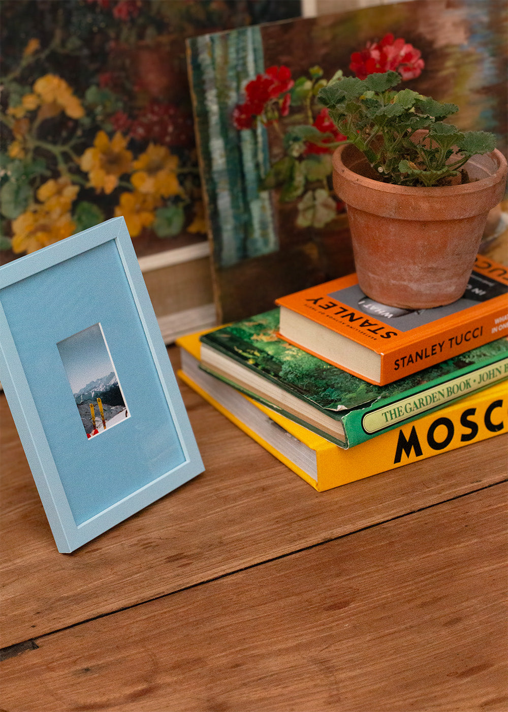 A light blue wooden picture frame with a small inset photograph, styled on a rustic wooden table next to a stack of books and a potted plant.