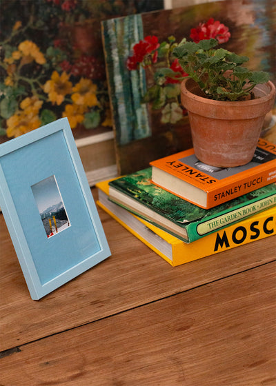 A light blue wooden picture frame with a small inset photograph, styled on a rustic wooden table next to a stack of books and a potted plant.