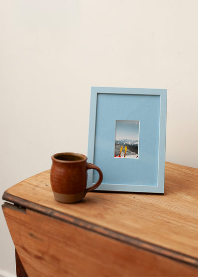 A light blue picture frame with a small landscape photograph inside, styled on a wooden table next to a brown ceramic mug against a plain off-white wall.