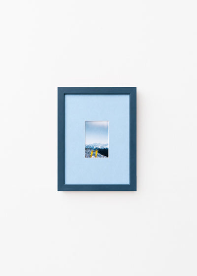 A dark blue picture frame with a light blue mat, displaying a small landscape photograph of mountains and yellow skis, set against a plain white wall.