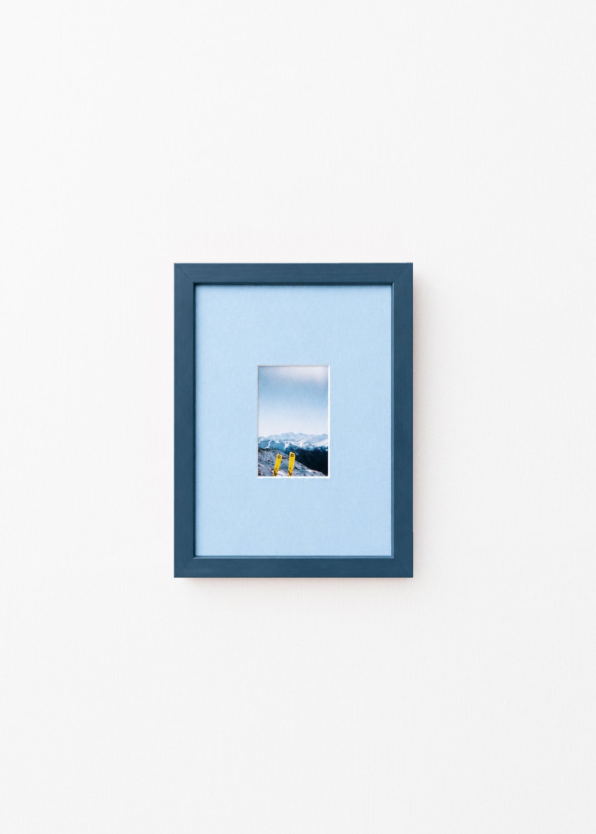 A dark blue picture frame with a light blue mat, displaying a small landscape photograph of mountains and yellow skis, set against a plain white wall.
