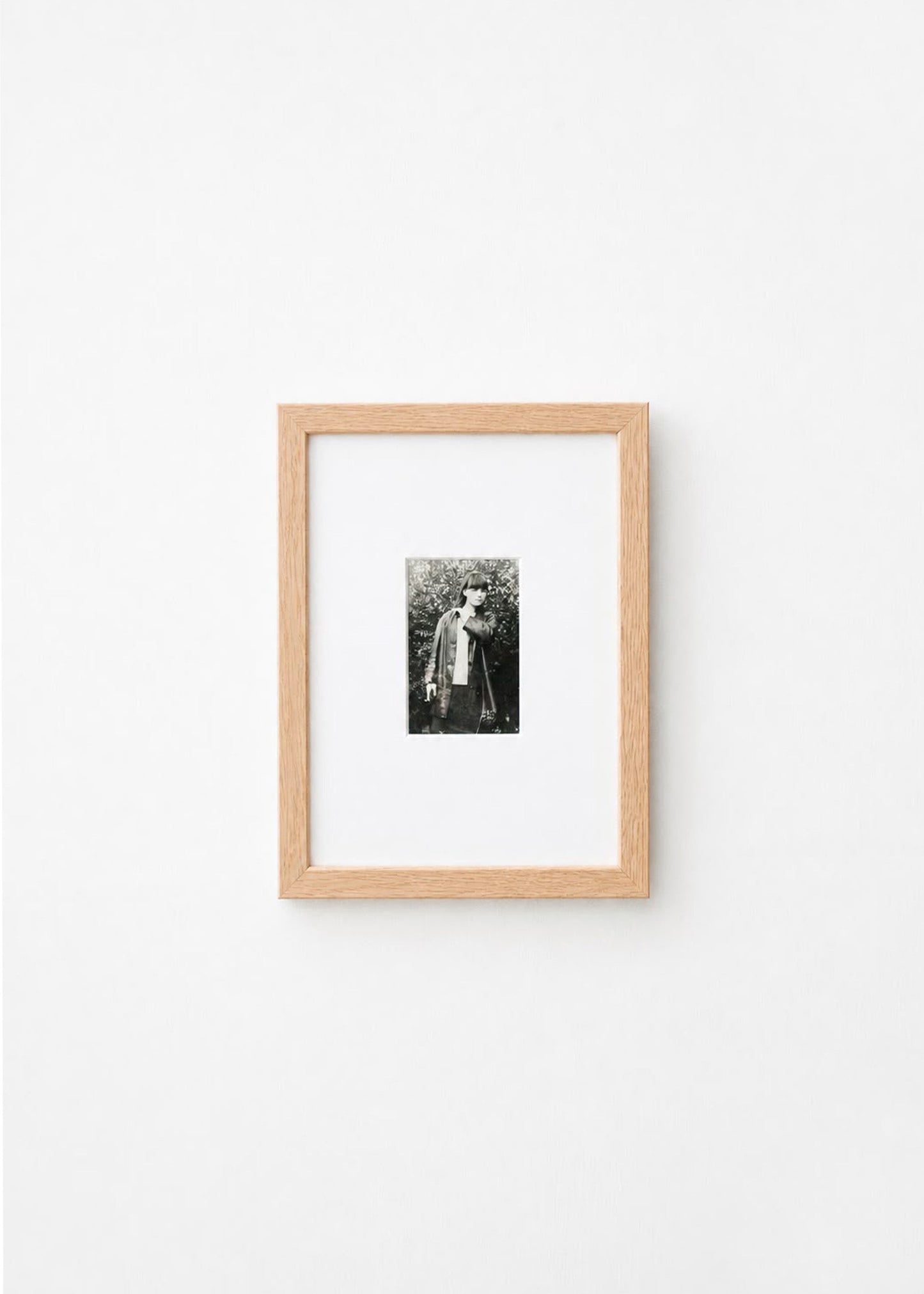 A small black and white portrait photograph in a light natural wood frame with a white mat, mounted on a plain white wall.
