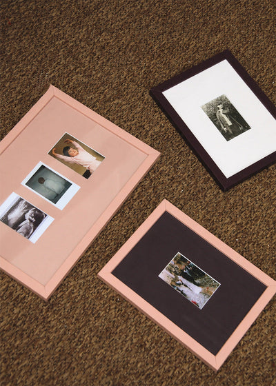 Three framed picture frames arranged on a brown carpeted floor. Two frames have light pink borders with white mats and small photos, and one frame has a dark brown border with a white mat and a larger photo.