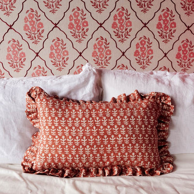 A decorative rectangular throw pillow with a ruffled edge, featuring a repeating white floral pattern on a terracotta background, styled on a bed with white pillows against patterned wallpaper.