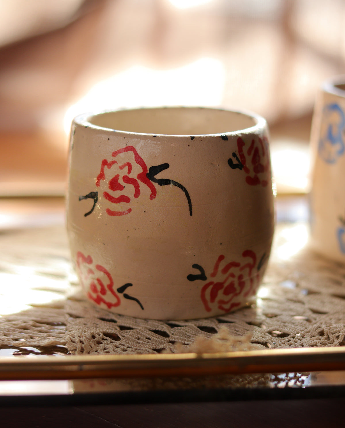 A beige ceramic stoneware mug with red floral pattern on it, placed on a lace tablecloth.