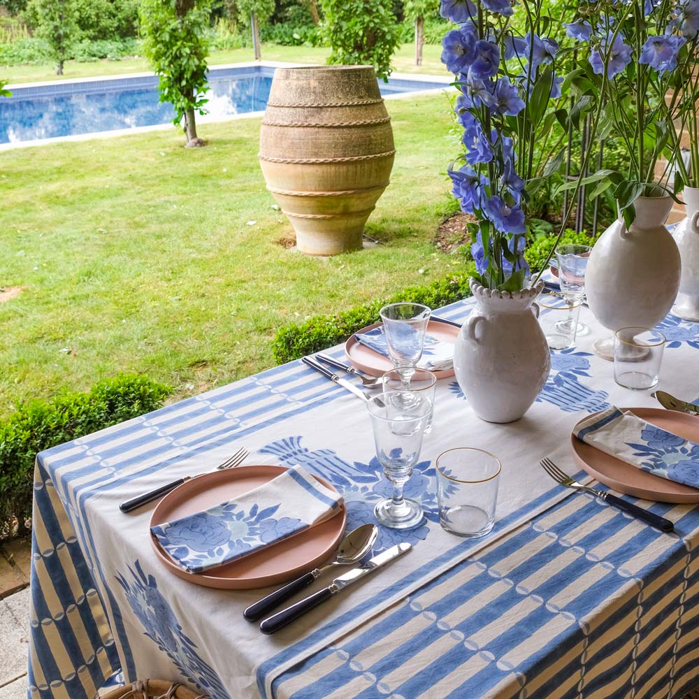 Table setting with Luna blue and white striped tablecloth, floral arrangements, and cutlery by a poolside.