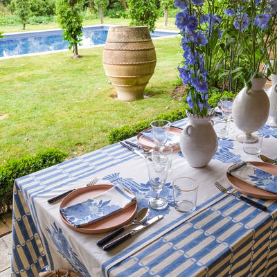 Table setting with Luna blue and white striped tablecloth, floral arrangements, and cutlery by a poolside.