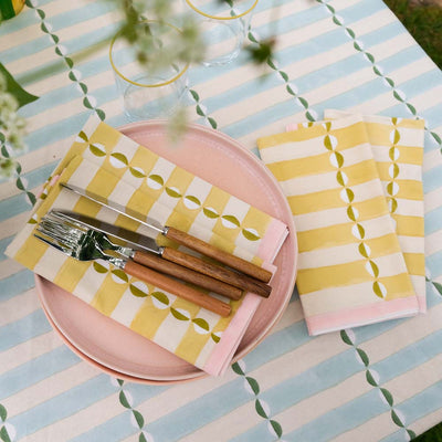 Table setting with pink plates, yellow and white patterned napkins, and wooden cutlery on a striped tablecloth.