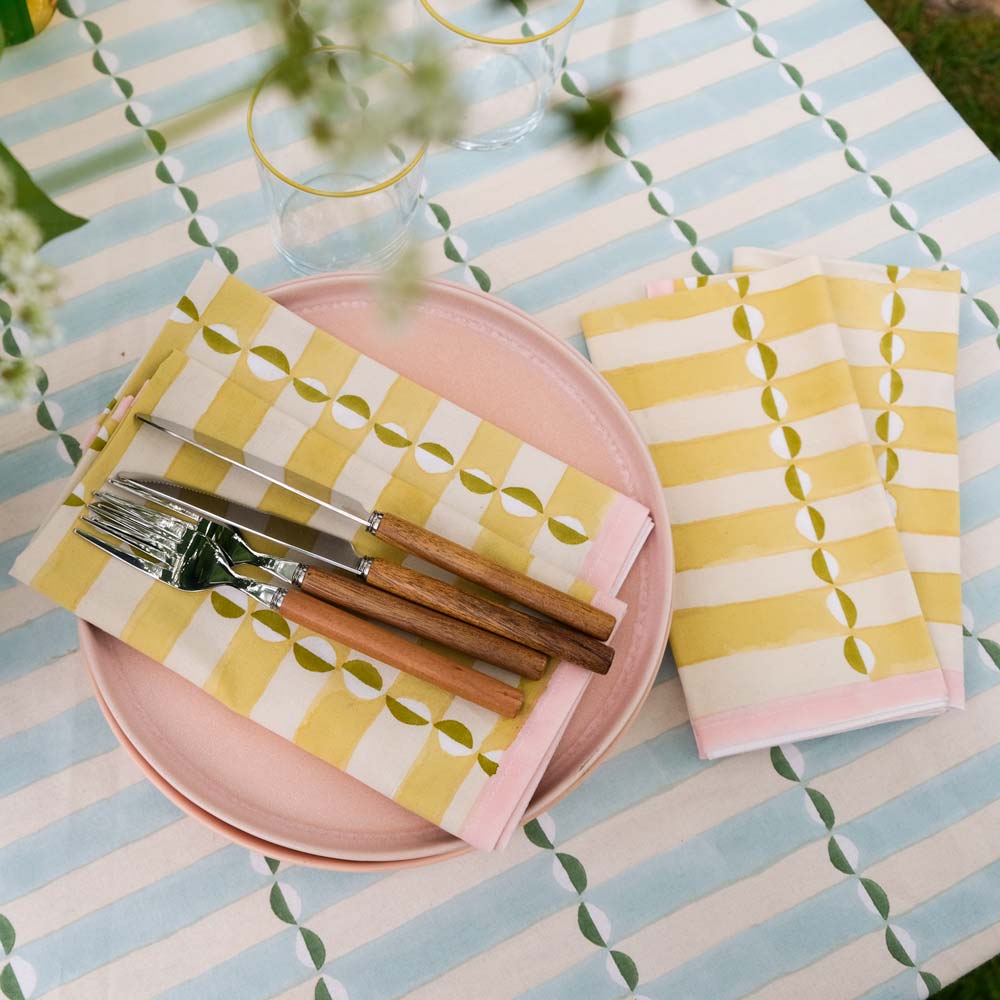 Table setting with pink plates, yellow and white patterned napkins, and wooden cutlery on a striped tablecloth.