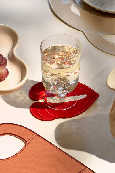 A glass of white wine rests on an Octaevo Red Anthurium Coaster beside plates and grapes on a cream tablecloth.